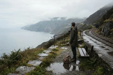 Lone traveler waiting at a rainy cliffside train stop above a misty coastal valley with curving railway tracks and distant sea cliffs.