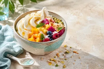 Creamy frozen yogurt topped with tropical fruit, toasted coconut, puffed grains, pistachios, basil leaves, and amber syrup on a bright stone table.