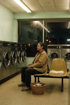 Solitary visitor sitting in a 1970s coin laundry at night beside chrome washing machines, mustard plastic chairs, and rain-streaked windows.