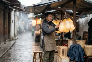 Young night market lantern keeper arranging glowing paper lanterns beneath a rainy canopy in an editorial photography scene.