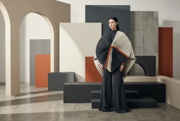 Fashion model in a sculptural pleated cape and wide-leg trousers standing among abstract geometric panels in a graphic gallery.