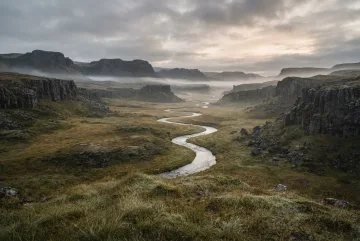 Fine art landscape photograph of a misty volcanic basalt valley with a winding silver stream, dark columnar rock formations, and golden alpine grasses under soft morning light.