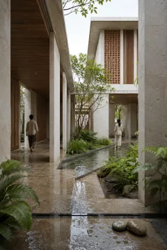 Portrait view of a rain-washed passive cooling garden pavilion with pale mineral walls, timber screens, terracotta ventilation blocks, reflective water channels, lush planting, and two anonymous visitors.