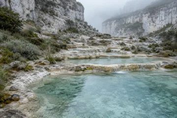 Rain-fresh pale limestone terraces descending into tranquil blue-green mineral lagoons beneath mist-softened cliffs and muted alpine vegetation.