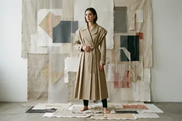 Fashion model in a sculptural stone-colored pleated trench coat standing among layered fabric, torn paper, acetate panels, and stitched textile shapes.
