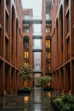Cinematic terracotta courtyard library with tall arches, rain-darkened stone paving, pedestrian bridges, mist, and warm interior lights.