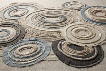 Abstract embroidered tidal rings made from layered linen and raised textile threads in clay, sand, dusty blue, charcoal, and ivory tones.